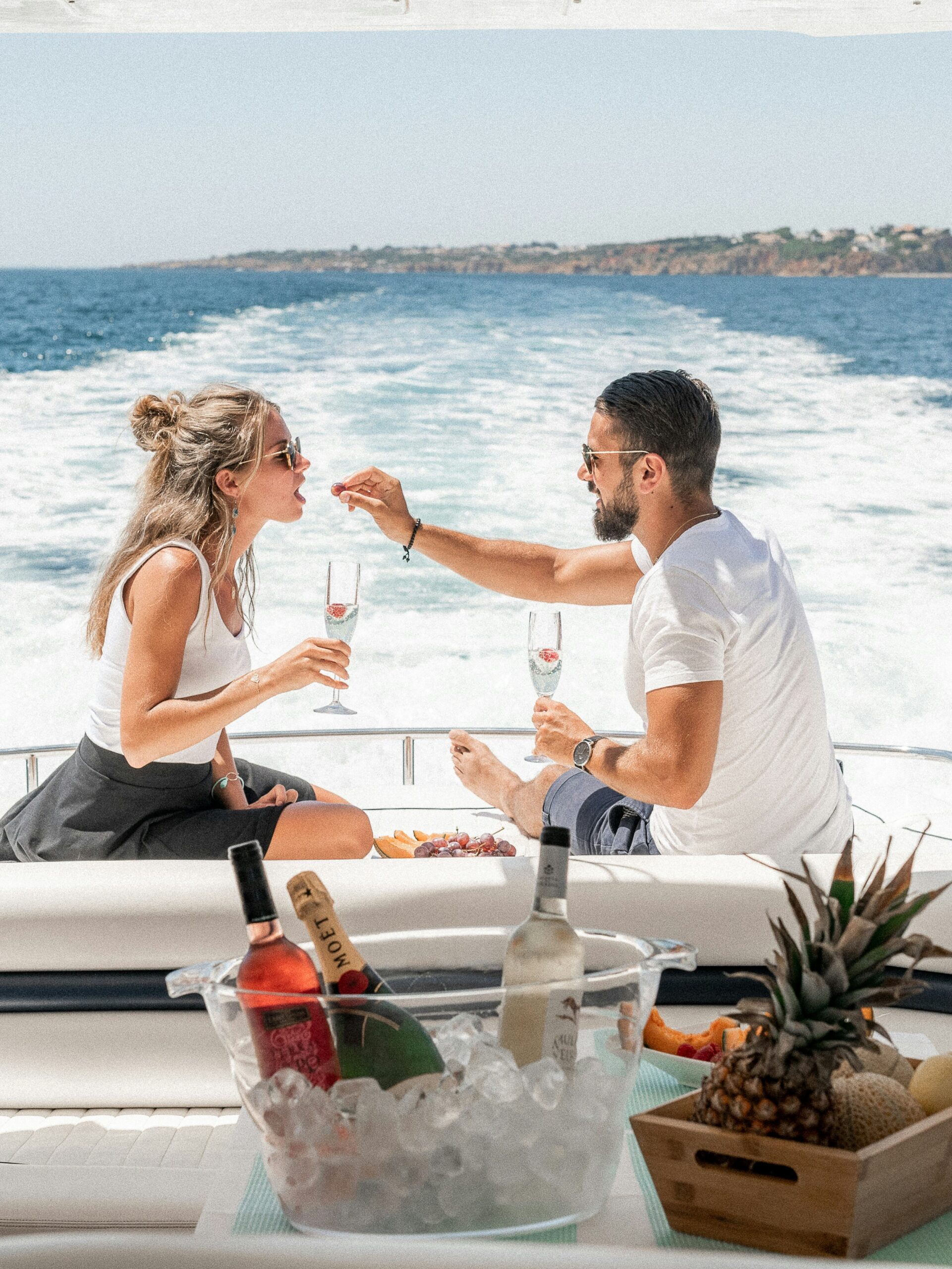 A couple enjoying a luxury experience on a boat, toasting with glasses of champagne, with a selection of fruits and drinks visible in an ice bucket nearby.