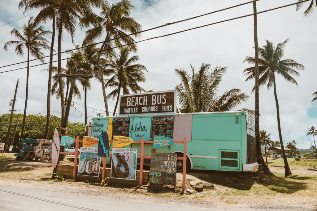 A colorful Hawaiian food truck serving waffles, churros, and fries, surrounded by palm trees and beach-themed signage.