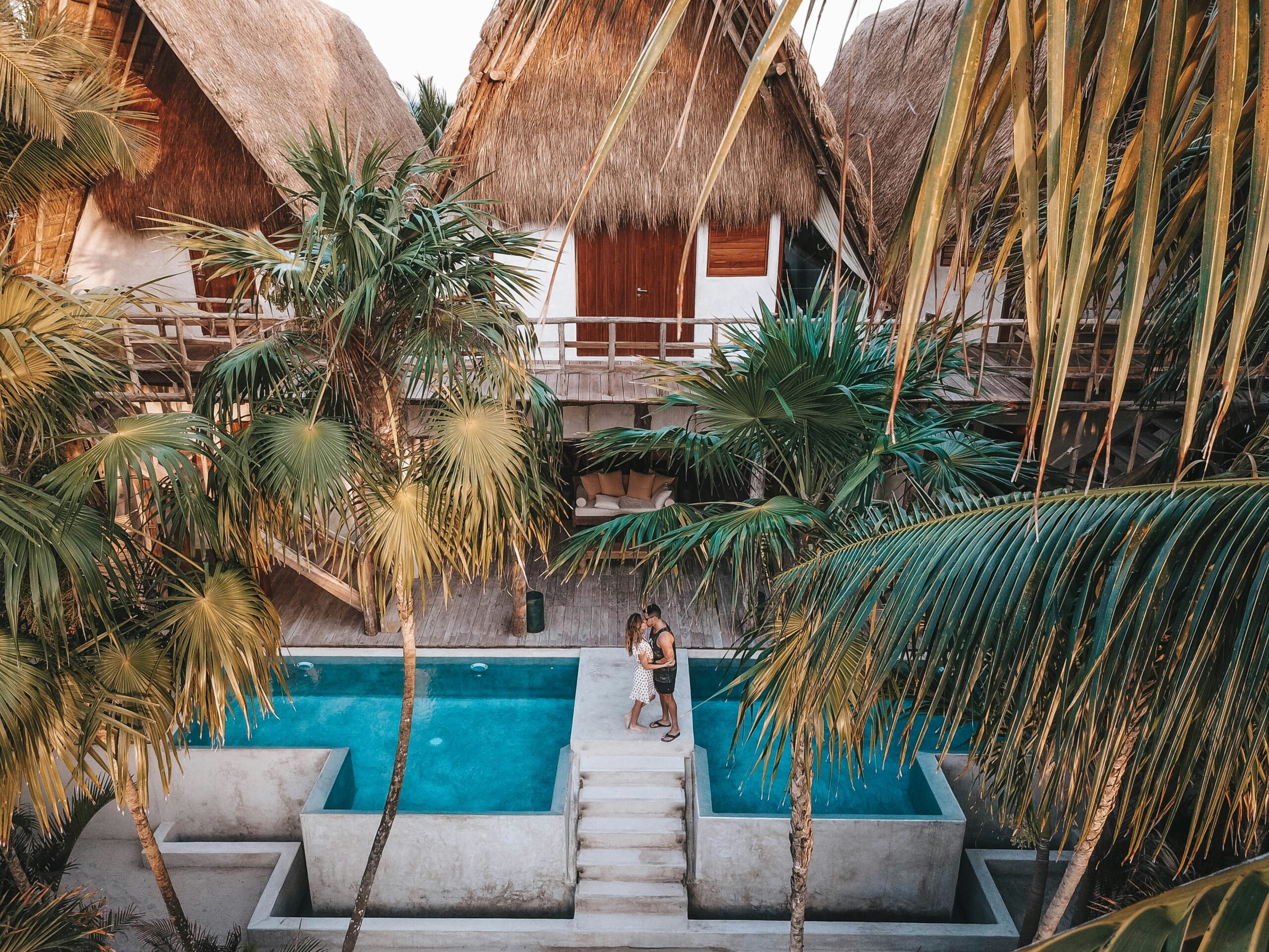 Couple embracing beside a tropical pool with palm trees and luxury thatched-roof villas in the background.